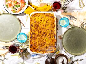 Pastel De Choclo in white dish, on a tabletop with plates, glasses of water and wine, and a salad on the side