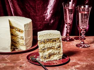 Alabama lane cake, with a slice placed on a red plate, on a red marble tabletop. 2 Magenta glasses of bubbly water, and a magenta fabric draped in the background.