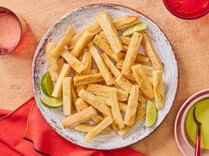 Fried yuca on a rustic plate, and textured terra cotta surface, with limes, dipping sauce and beverage on the side