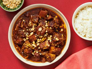 Overhead of stew in a bowl with small dish of peanuts in bowl to the side with red napkin and red background