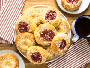 Kolaches on a platter, with one cream and one strawberry on 2 plates, with coffee on the side, striped napkins, on a wooden surface.