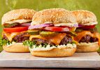 Three cheeseburgers with lettuce, tomato, pickle, onion, and cheddar cheese on a sesame seed bun displayed on a cutting board