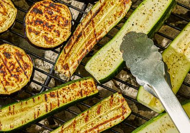 Slices of zucchini and eggplant grilling on a barbecue with metal tongs