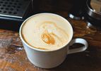 A cup of coffee on a wooden table with foam art on the surface, a coffee machine partially visible in the background