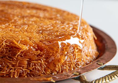 Knafeh being drizzled with syrup from above set on a round copper tray with handles