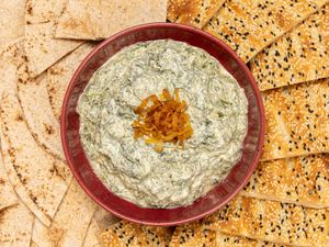 A plate of creamy spinach dip garnished with fried onions surrounded by flatbread and sesame crackers