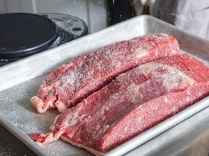 Salt being sprinkled on two raw beef cuts on a tray