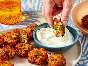 A plate of fritters with a hand dipping one into a yogurtbased sauce a glass in the background on a patterned surface