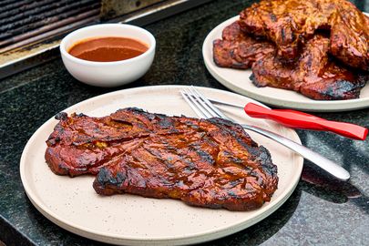 Cooked pork steak on a plate with barbecue sauce and utensils additional steaks in the background on a platter on a black countertop