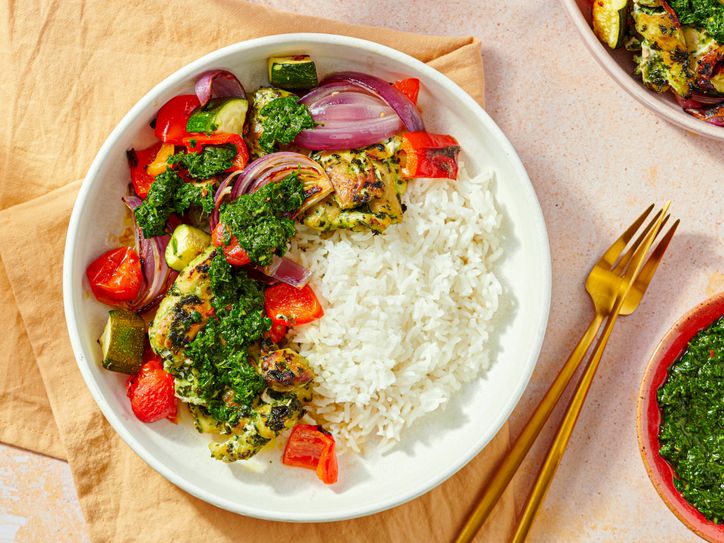 A dish with grilled chicken thighs vegetables chimichurri sauce and white rice on a plate utensils and a sauce bowl beside it